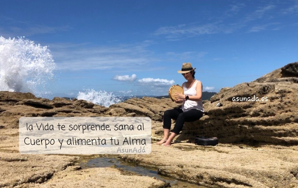 Asun Adá sentada en una roca junto al mar, con sombrero y tocando la pandereta con caja de texto: la Vida te sorprende, sana al Cuerpo y alimenta tu Alma, por AsunAdá 